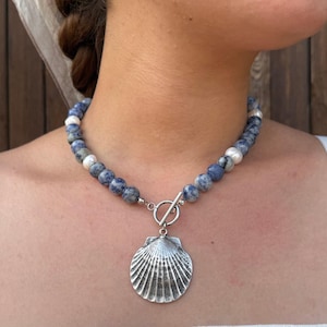 May include: A beaded necklace featuring round blue and white mottled stone beads interspersed with small white pearls. The necklace includes a silver-toned metal toggle clasp at the front, from which hangs a large, textured silver-toned seashell pendant.