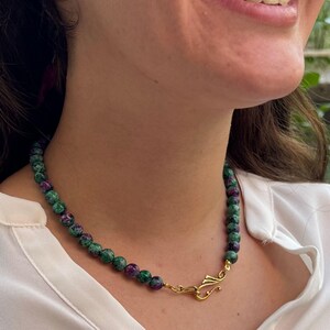 May include: A beaded necklace featuring round, mottled green and deep magenta gemstone beads strung together. The necklace is secured with a gold-toned metal hook-and-eye clasp positioned at the front, connecting the two ends of the beaded strand.
