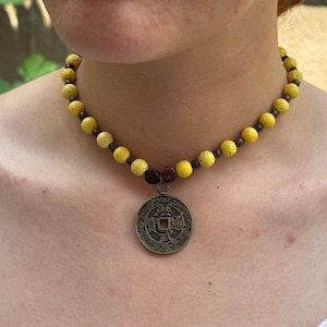May include: A yellow beaded necklace with a bronze coin pendant. The necklace features round, textured yellow beads and smaller brown beads. The coin pendant has an intricate design. The necklace is worn around the neck.