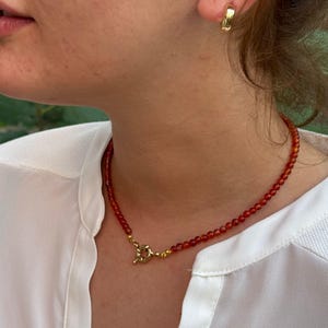 May include: A beaded necklace featuring small, round, deep orange-red carnelian stones strung together, finished with a gold-toned metal toggle clasp at the front. A matching gold-toned hoop earring is visible on the earlobe.