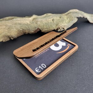 Peut inclure: Un porte-cartes en bois brun avec un couvercle articul&eacute;, r&eacute;v&eacute;lant une carte cadeau. La carte cadeau affiche le logo Amazon et le texte "&euro;10". Le support est en bois stratifi&eacute; et pr&eacute;sente un aspect textur&eacute;.