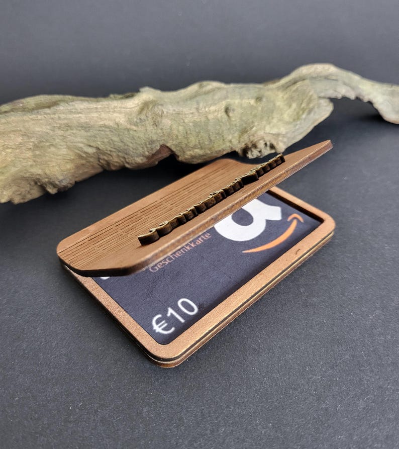 Peut inclure: Un porte-cartes en bois brun avec un couvercle articul&eacute;, r&eacute;v&eacute;lant une carte cadeau. La carte cadeau affiche le logo Amazon et le texte "&euro;10". Le support est en bois stratifi&eacute; et pr&eacute;sente un aspect textur&eacute;.