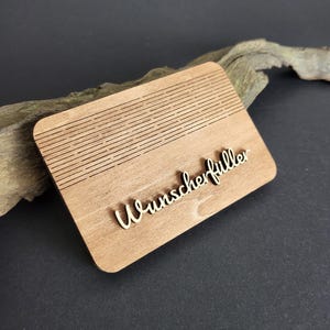 Peut inclure: Plaque rectangulaire en bois clair avec le mot "Wunscherf&uuml;ller" en &eacute;criture cursive. La partie sup&eacute;rieure de la plaque pr&eacute;sente des lignes horizontales grav&eacute;es. La plaque est pos&eacute;e sur une surface sombre.