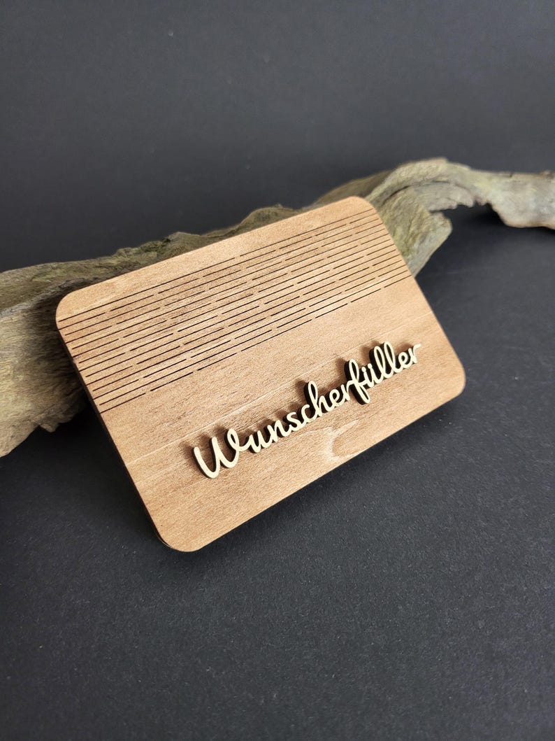 Peut inclure: Plaque rectangulaire en bois clair avec le mot "Wunscherf&uuml;ller" en &eacute;criture cursive. La partie sup&eacute;rieure de la plaque pr&eacute;sente des lignes horizontales grav&eacute;es. La plaque est pos&eacute;e sur une surface sombre.