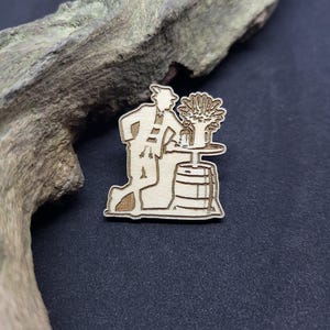 Könnte beinhalten: Ein Holz-Anstecknadel mit einer stilisierten Illustration einer Person mit Hut und Mantel, die sich in der Nähe eines Fasses und eines Tisches mit einem Blumenarrangement kniet. Die Anstecknadel ist aus hellem Holz gefertigt und hat eine rustikale Ästhetik.
