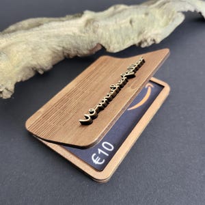 Peut inclure: Un porte-cartes en bois avec un couvercle &agrave; charni&egrave;re, orn&eacute; d'une &eacute;criture d&eacute;corative et d'une carte cadeau visible &agrave; l'int&eacute;rieur. La carte cadeau affiche le logo Amazon et la valeur de 10 &euro;.