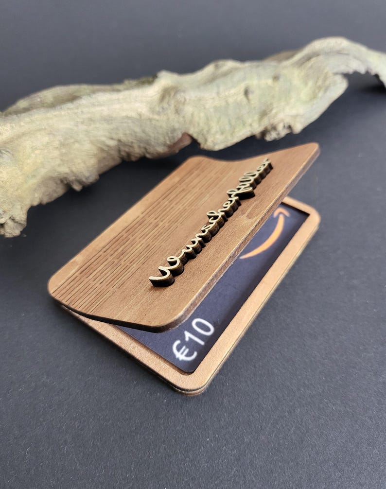 Peut inclure: Un porte-cartes en bois avec un couvercle &agrave; charni&egrave;re, orn&eacute; d'une &eacute;criture d&eacute;corative et d'une carte cadeau visible &agrave; l'int&eacute;rieur. La carte cadeau affiche le logo Amazon et la valeur de 10 &euro;.