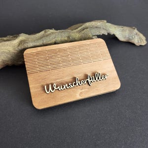 Peut inclure: Plaque rectangulaire en bois clair avec le mot "Wunscherf&uuml;ller" en &eacute;criture cursive. La plaque pr&eacute;sente des lignes horizontales grav&eacute;es dans la partie sup&eacute;rieure. Fond noir avec un morceau de bois flott&eacute;.