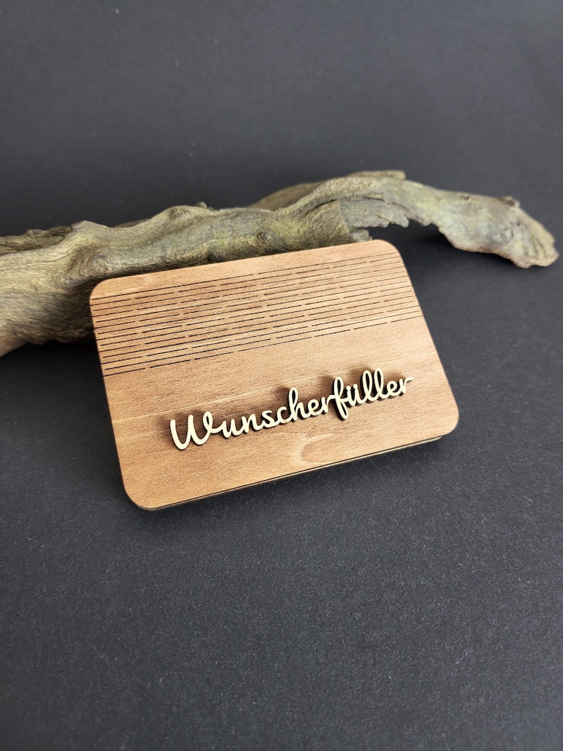 Peut inclure: Plaque rectangulaire en bois clair avec le mot "Wunscherf&uuml;ller" en &eacute;criture cursive. La plaque pr&eacute;sente des lignes horizontales grav&eacute;es dans la partie sup&eacute;rieure. Fond noir avec un morceau de bois flott&eacute;.