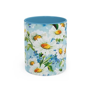 May include: A white ceramic mug with a blue rim and a floral pattern of white daisies and blue forget-me-nots on a light blue background.