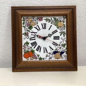 May include: A square wall clock with a dark brown wooden frame and a white ceramic face. The clock features black Roman numerals and a decorative border of colorful painted fruits, including oranges, grapes, and pears, with green leaves.