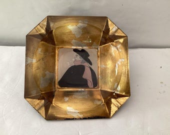 Cenicero vintage con retrato estilizado de un hombre con sombrero negro y bufanda rosa, del siglo XIX, con detalles dorados.