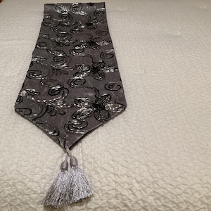 May include: A gray table runner with a silver floral pattern and tassels. The runner is made of a soft, velvety fabric.