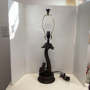 May include: A bronze-colored table lamp with a decorative base featuring a seated figure. The lamp has a curved stem, a floral accent, and a brass-colored lamp holder. The lamp is approximately 24 inches tall.