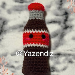 May include: A crocheted plush toy shaped like a soda bottle with a red and white striped label and a cute face. The toy has black eyes and a pink mouth. The text "@Yazendiz" is visible on the toy.