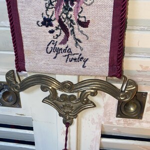 Closeup of signature area on Glenda Turley woven textile wall hanging showing artist name integrated into tapestry design along with lower floral ribbon and bird motif ornate brass style hardware and tassel attachment point highlighting authenticity fiber art detail vintage decorative wall hanging craftsmanship