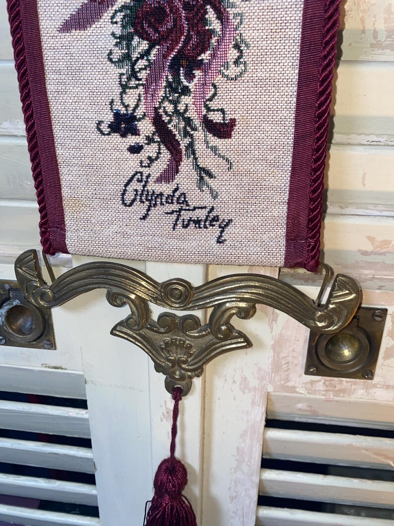 Closeup of signature area on Glenda Turley woven textile wall hanging showing artist name integrated into tapestry design along with lower floral ribbon and bird motif ornate brass style hardware and tassel attachment point highlighting authenticity fiber art detail vintage decorative wall hanging craftsmanship