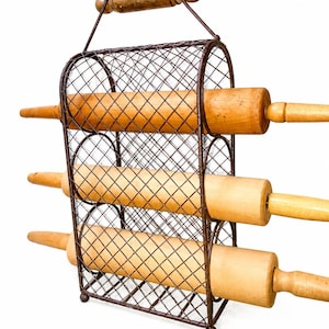 May include: A brown metal wire rack with a diamond-patterned mesh frame and a wooden handle at the top. The rack holds three light-colored wooden rolling pins, each positioned horizontally within the three circular storage sections of the frame.