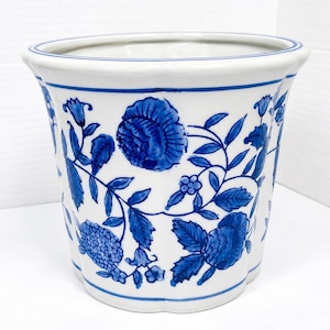 May include: A white ceramic planter featuring a hand-painted blue floral pattern with detailed blossoms, leaves, and vines. The vessel has a scalloped rim and a wide, tapered cylindrical shape with a smooth, glossy finish throughout the exterior.