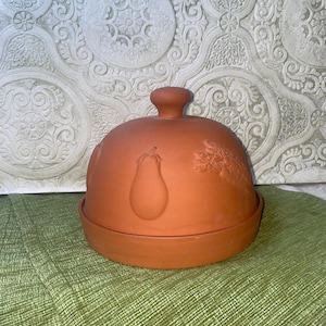 May include: A terracotta cloche with a round base and a domed lid. The lid features embossed images of a pear and wheat. The cloche is designed for food storage and presentation. It sits on a green textured surface.