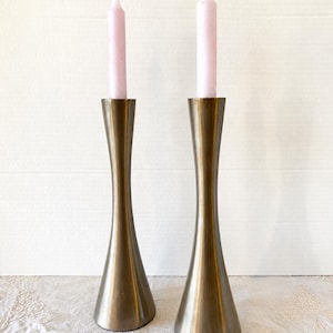 May include: Two brass-toned metal candle holders with a flared base and a slender, hourglass-shaped body. Each holder supports a pale pink candle. The candle holders are displayed on a white surface.