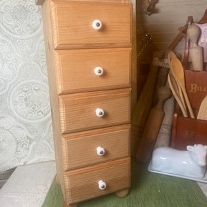 May include: A small, light-colored wooden cabinet with five drawers. Each drawer has a white knob with a decorative edge. The cabinet is rectangular and sits on four small, rounded feet. The wood grain is visible.