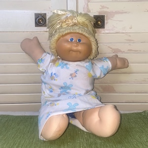 May include: A vintage Cabbage Patch Kid doll with blonde yarn hair and blue eyes. The doll is wearing a white dress with blue floral and bunny print. The doll is sitting on a green rug.