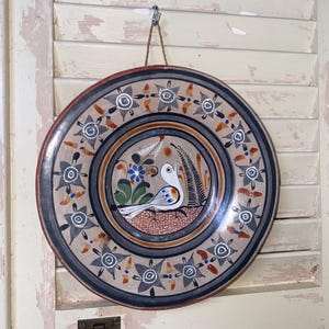 May include: A decorative ceramic plate with a hand-painted bird design. The plate features a central image of a white bird with blue and orange accents, surrounded by floral and geometric patterns in shades of gray, brown, and blue. The rim is a dark red.