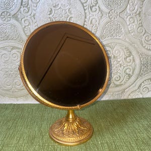May include: A vintage, round vanity mirror with a gold-colored metal frame and base. The mirror is set on a decorative pedestal with ornate detailing. The mirror's surface reflects a dark image. The base is approximately 4 inches in diameter.