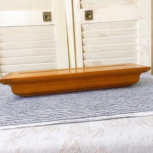 May include: A rectangular wooden display base with a smooth, medium-brown finish and a long, narrow slot running horizontally across the top surface. The object features beveled edges and a slightly curved profile along the sides.