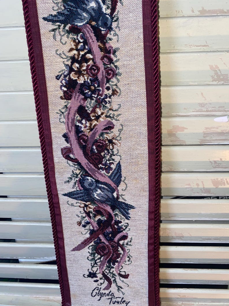 Op de afbeelding: Een decoratief tapijt met een vogel- en bloemenontwerp. Het tapijt heeft een centraal patroon van een blauwe vogel, bordeauxrode linten en diverse bloemen op een beige achtergrond. De randen zijn afgewerkt met een bordeauxrood touw. De tekst "Glynda Turkey" staat onderaan.