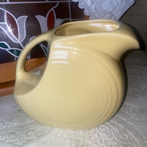 May include: A pale yellow ceramic pitcher with a curved handle and spout. The pitcher has a rounded body with concentric ridges and a smooth, glossy finish. The pitcher is sitting on a patterned surface.