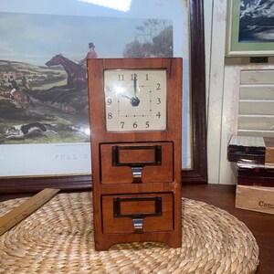 May include: A wooden desk clock with a square clock face and two small drawers. The clock face has black numerals and hands. The drawers have metal label holders and pulls. The clock is set on a woven mat.