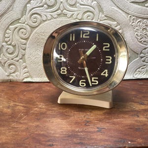 May include: A vintage Baby Ben alarm clock with a gold-tone bezel and a dark brown face. The clock features large, easy-to-read white numerals and hands. The clock is mounted on a cream-colored base.