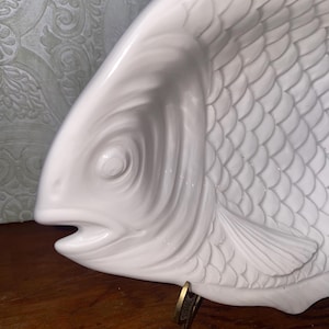 May include: A white ceramic fish-shaped serving dish. The dish features detailed scales and fins, with an open mouth. The dish is designed to sit on a flat surface. The overall design is a decorative piece for serving food.