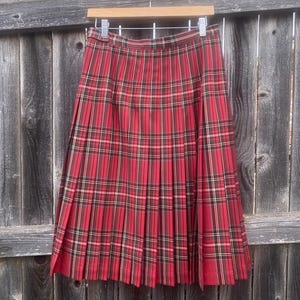 May include: A pleated red plaid skirt with a classic pattern. The skirt features a traditional tartan design with red, black, and white lines. It hangs on a wooden hanger against a weathered wooden fence.
