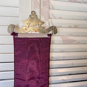 Op de afbeelding: Decoratieve wandophanging met een messingkleurige metalen beugel met een gestileerd ontwerp. Een diep bordeauxrood stoffen paneel met een getextureerd oppervlak en een gevlochten rand hangt eronder. De achtergrond is een witte shutter.