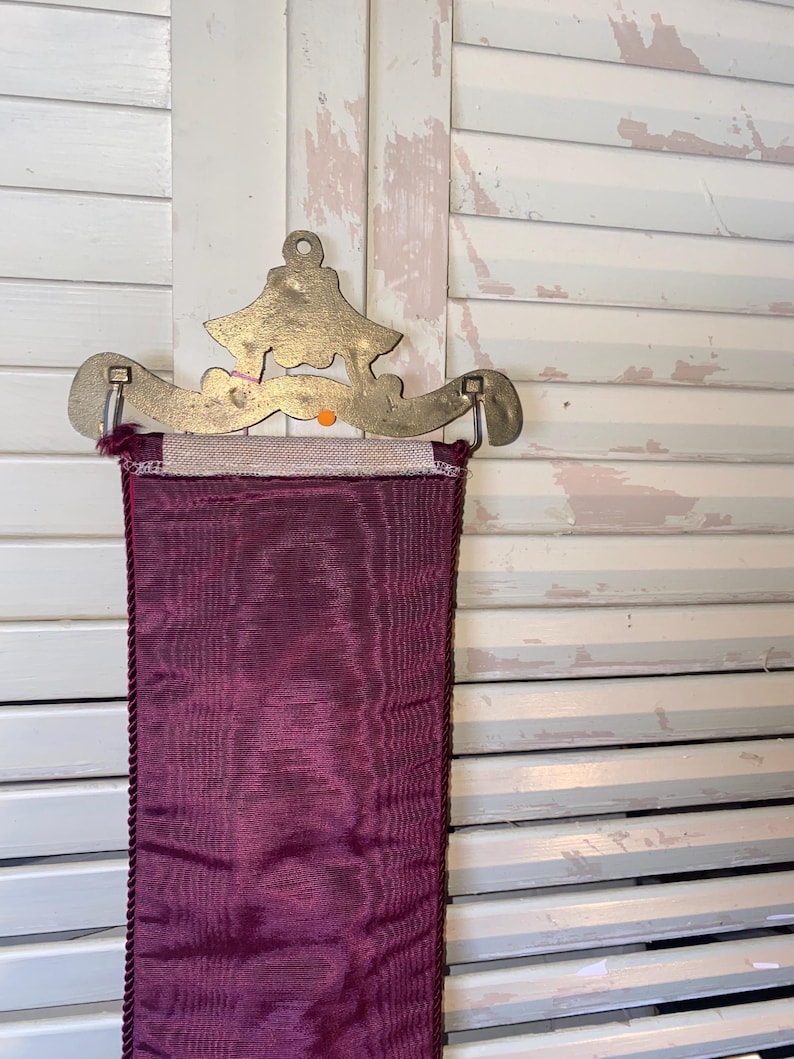 Op de afbeelding: Decoratieve wandophanging met een messingkleurige metalen beugel met een gestileerd ontwerp. Een diep bordeauxrood stoffen paneel met een getextureerd oppervlak en een gevlochten rand hangt eronder. De achtergrond is een witte shutter.