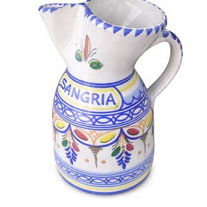 May include: White ceramic sangria pitcher with a unique spout design. The pitcher is decorated with blue, yellow, red, and green patterns, and the word "SANGRIA" is prominently displayed in a blue and yellow oval.