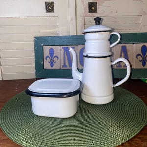 May include: A white enamel coffee pot with a black handle and lid, and a matching square container with a black rim. The items are on a green placemat, with a decorative sign in the background.