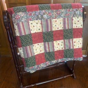 May include: A patchwork quilt with red, green, and cream-colored squares, adorned with star and floral patterns, is displayed on a dark wooden quilt rack. The quilt rack has a curved top and a horizontal bar at the bottom.