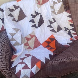 May include: A handmade quilt with a white background and geometric patterns in shades of brown, orange, and beige. The quilt is draped over a wicker chair, showcasing its intricate design and craftsmanship. The quilt's dimensions are not visible.