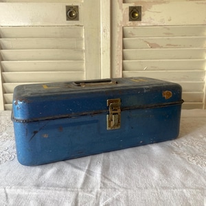 May include: A vintage blue metal toolbox with a handle and a brass clasp. The toolbox has a rectangular shape and shows signs of wear, with some yellow lettering on the top. The background features white shutters and a white tablecloth.