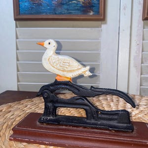 May include: A vintage duck-shaped nutcracker. The duck is white with orange accents and sits atop a black metal nutcracker mechanism. The base is a dark brown wooden rectangle. The background includes a painting and white shutters.