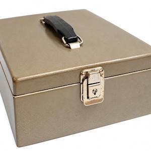May include: A rectangular metal storage box in a muted taupe finish. The box features a black plastic carrying handle mounted on the top lid and a silver-toned metal latch mechanism on the front center, which includes a small integrated keyhole.