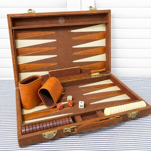 May include: A brown backgammon set in a briefcase-style case with a brown felt interior, featuring cream and brown triangular points. The set includes two brown leather dice cups, brown and cream checkers, two red dice, and a white doubling cube with 4 and 32.