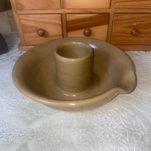 May include: A tan-colored ceramic candle holder with a circular base and a central cylindrical cup. The holder has a small spout on the side. The background includes a wooden cabinet with drawers.