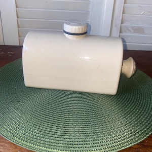May include: A rectangular, off-white ceramic hot water bottle with a rounded top and a small, circular stopper. The bottle has a rounded handle on one end. The stopper is white with a black band. The bottle is on a green placemat.