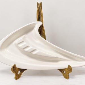 May include: A glossy off-white ceramic dish with an elongated, asymmetrical crescent shape and a series of raised, stepped ridges along the center. The dish is displayed on a decorative, textured gold-toned metal stand.