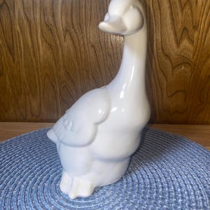 May include: A white ceramic goose figurine with a long neck and detailed feathers. The goose is standing upright, with its head turned slightly to the side. The figurine is on a blue placemat.